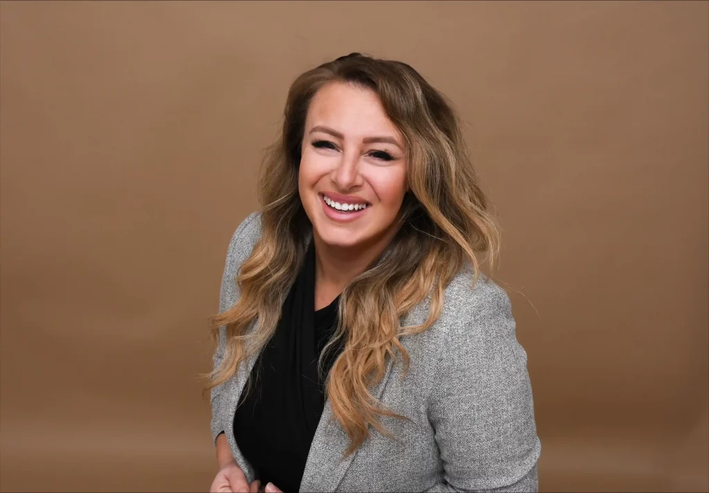 Das Bild fängt einen freudigen Moment einer Frau mit langem, welligem Haar ein, das in sanften Tönen gestylt ist. Sie lächelt breit und strahlt Wärme und Selbstvertrauen aus, während sie einen eleganten grauen Blazer über einem schwarzen Oberteil trägt. Der Hintergrund ist in einem dezenten Braun gehalten, das ihre Kleidung ergänzt und ihren fröhlichen Ausdruck hervorhebt.